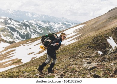 Charming girl tourist with a large backpack in sunglasses walks along the slope of a snow-covered mountain on a warm spring day. Concept of travel and new experiences
