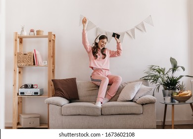 Charming girl in tracksuit dancing, sitting on couch. Woman holding smartphone and listening to music with headphones. Shot of teen in pink home suit having fun in cozy home atmosphere