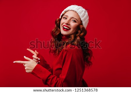 Charming girl with wavy hair expressing happiness in christmas. Pretty female model in knitted hat laughing on red background.