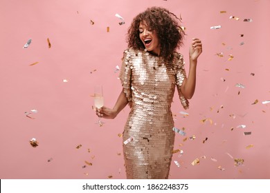 Charmantes Mädchen mit scharfen kurzen Haaren in glänzendem beigem Kleid, das Glas mit Champagner hält und mit Konfetti auf rosafarbenem Hintergrund posiert.