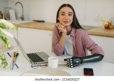 Charming gorgeous adorable photogenic woman with full pouty lips and iron bionic arm prosthesis, holding pen in hand, looking at camera, working at kitchen table, making notes next to laptop and phone