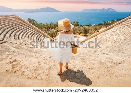 Charming and gorgeous woman in white dress and hat explores ancient landmark and ruins of Greek or Roman amphitheater on the resort coast of the Mediterranean Sea