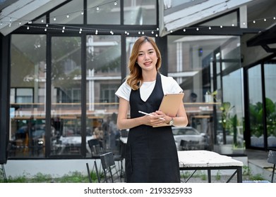 Charming And Gorgeous Young Asian Female Restaurant Or Coffee Shop Entrepreneur Standing In Front Of The Entrance With A Smile On Her Face.