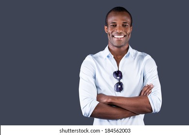 Charming Handsome. Handsome Young Black Man In Blue Shirt Keeping Arms Crossed And Smiling At Camera While Standing Against Grey Background