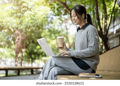 A charming, happy Asian woman enjoys a hot drink while working remotely outdoors, sits on a park bench with her laptop, wearing earphones. - Powered by Shutterstock