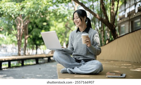 A charming, happy Asian woman enjoys a hot drink while working remotely outdoors, sits on a park bench with her laptop, wearing earphones. - Powered by Shutterstock