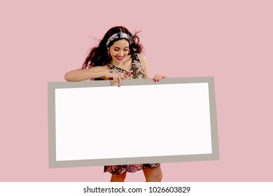 Charming And Happy Freshing Face Woman With Wind Blowing Hair.Holding Billboard / Blank Sign Looking Down. Isolated On Pink Background