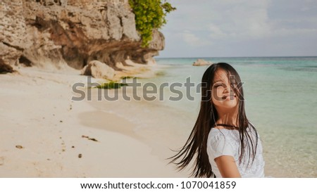 A charming and happy philippine teenage girl in a white summer dress is running along a tropical beach near the rocks. She is happily spinning. Childhood. Recreation.