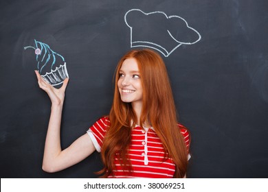 Charming Happy Redhead Young Woman In Drawn Chef Hat Holding Drawing Cupcake Over Blackboard Background