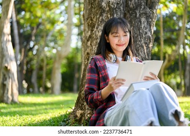 Charming and happy young Asian woman reading a book or novel under the tree in the beautiful green park. - Powered by Shutterstock