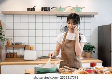 A charming, happy young Asian woman in an apron is talking on the phone while cooking in the kitchen, frying something in a pan. lifestyles, home cooking, food - Powered by Shutterstock