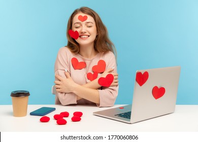 Charming happy young woman with kind smile sitting at workplace office, all covered with sticker love hearts, embracing herself, enjoying valentine's day greetings. indoor studio shot, isolated