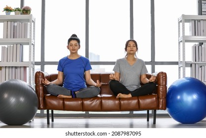 Charming and healthy Asian woman sitting cross legged camly, eye close, deep breathing on couch for easy yoga meditation pose as Sukhasana practice for peaceful mind, stress relief in living room.