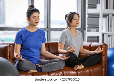 Charming and healthy Asian woman sitting cross legged camly, eye close, deep breathing on couch for easy yoga meditation pose as Sukhasana practice for peaceful mind, stress relief in living room.