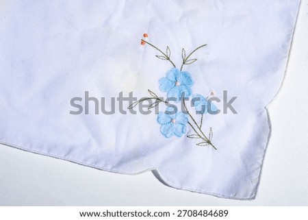charming, high-resolution studio shot of white fabric, likely a handkerchief, adorned with delicate blue floral embroidery, showcasing intricate detail and a scalloped edge.
