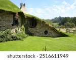 A charming hobbit-style cottage with a grass-covered roof nestled in a lush rural landscape, featuring round windows and stone walls.