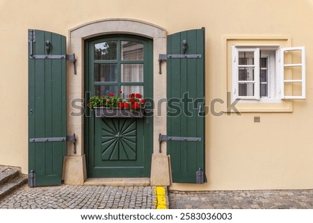 A charming house facade features a vibrant green door with decorative carvings, matching shutters, a window box of red flowers, and an adjacent white window against a pale yellow wall.