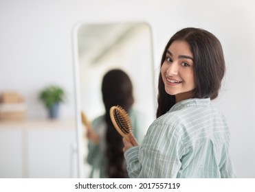 Charming Indian woman with long beautiful hair standing with wooden brush in front of mirror, looking at camera over her shoulder at home. Young lady taking care of herself. Hairdressing and styling
