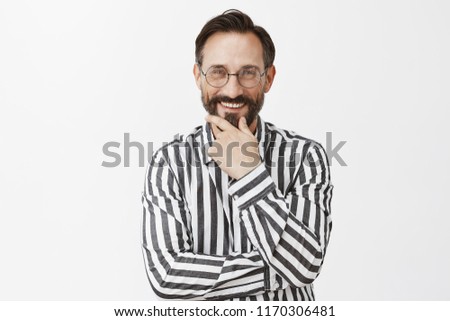 Charming and joyful adult male model with moustache in glasses and striped shirt, touching beard and smiling broadly while casually talking with friend, having funny and amusing talk over grey wall