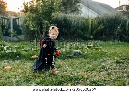 A charming kid in a devil costume and a raincoat sneaks around the garden. Halloween concept, carnival costume
