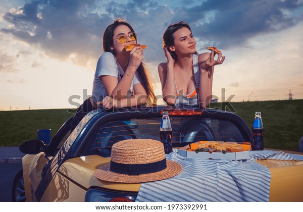 Charming ladies in casual outfit are eating\
pizza, posing in yellow car with french fries, hat and soda in\
glass bottles on trunk. Fast food. Mock\
up