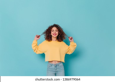 Charming lady in yellow sweater and skinny jeans smiles and touches her curly dark hair against blue background