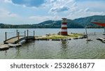 Charming lighthouse on a small island connected by a wooden bridge on lake Stubenbergsee surrounded by rolling hills of East Styrian Hill Country in Styria, Austria. Lush greenery in the background