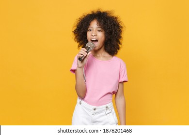 Charming Little African American Kid Girl 12-13 Years Old In Pink T-shirt Isolated On Yellow Wall Background Studio Portrait. Childhood Lifestyle Concept. Mock Up Copy Space. Sing Song In Microphone