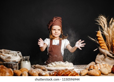 Charming little girl in apron and hat standing at table kneading bread dough and having fun spending time cooking
