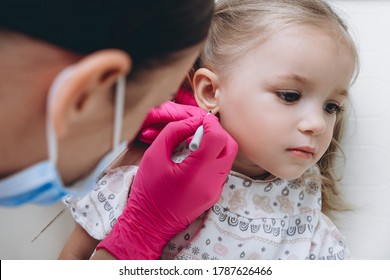 Charming little girl having an ear piercing process with special equipment in a beauty center by a professional medical worker