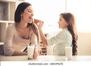 Charming little girl and her beautiful young mom are eating muffins with milk and smiling while resting at home