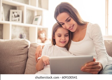 Charming Little Girl And Her Beautiful Young Mom Are Using A Digital Tablet And Smiling While Sitting On Sofa At Home