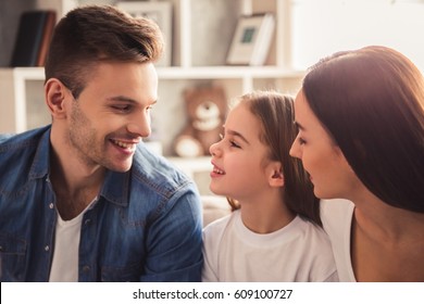 Charming little girl and her beautiful young parents are talking and smiling while sitting on sofa at home
