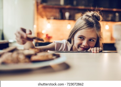 Charming little girl on kitchen is looking from under the table on sweets. Ready to eat some cookies and cakes.