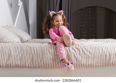 Una encantadora niñita de pajama rosa con bata está sentada en la cama, poniéndose sus zapatillas suaves y sonriendo.