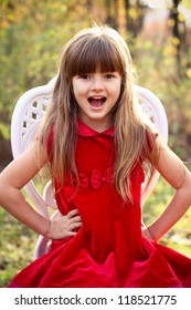 Charming Little Girl In A Red Dress Sitting On A Chair In The Autumn Forest