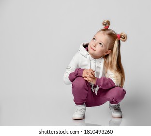 Charming Little Girl With A Charming Smile Sitting On The Floor In A Trendy Sweat Suit On A Gray Background. The Girl's Upward Gaze Is Focused On The Free Space For Text Or Promotional Items.