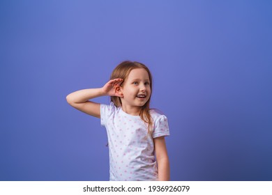 Charming Little Girl Smiles With Her Hand Over Her Ear While Listening And Hearing Rumors Or Gossip On A Purple Isolated Background. Child Eavesdropping