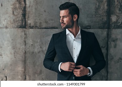 Charming Man. Handsome Young Man In Suit Looking Away And Adjusting His Jacket While Standing Indoors   