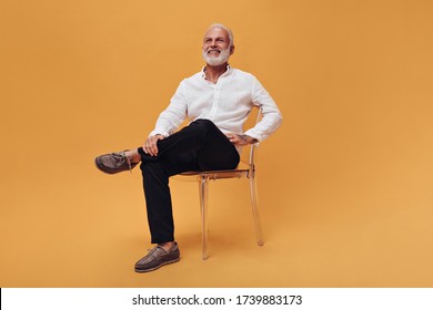 Charming man in white shirt and black pants sitting on orange background. Handsome adult guy in good mood is sitting on chair - Powered by Shutterstock