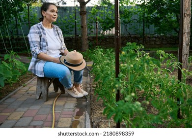 Charming Mature Hispanic Woman Gardener, Sitting On A Wooden Stool And Resting After Hard Work In Her Own Ecological Farm. Agricultural Hobby, Cultivation Organic Vegetables, Horticulture, Gardening