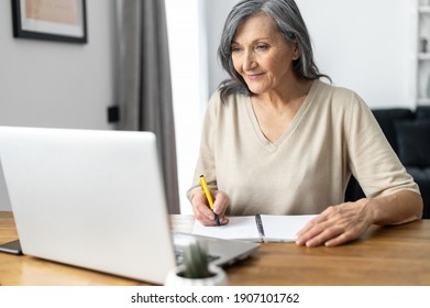 Charming Mature Mid-aged Woman Watching Online Webinars On The Laptop Sitting At The Table At Home, Taking Notes, E-learning. Senior Business Lady Researching, Analyzing Tasks Works In The Home Office