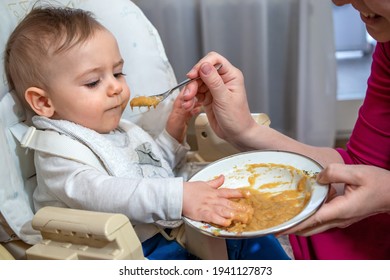Charming Messy Little Baby Boy Sitting In A Feeding Chair, Eating And Playing With His First Food From The Spoon With Mom At Home. 
