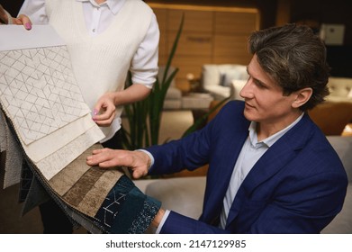 Charming Middle Aged Man, Interior Designer In Business Suit Puzzled By The Choice Of Upholstery Material For A New Sofa, Examines Fabric Samples In The Hands Of A Sales Assistant In A Furniture Store