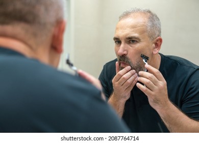 Charming middle aged man shaving his beard and moustache off in front of a mirror to look younger