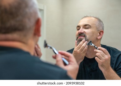 Charming middle aged man shaving his beard and moustache off in front of a mirror to look younger