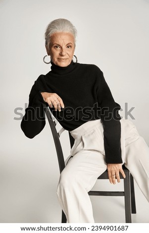 charming middle aged woman in smart casual outfit and hoop earrings sitting on chair on grey