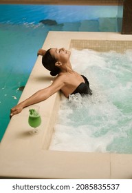 Charming middle-aged brunette, gorgeous woman enjoying relaxing time lying with closed eyes in a thermal pool, getting hydromassage in wellness spa resort. Purity, hydrotherapy and body care concept