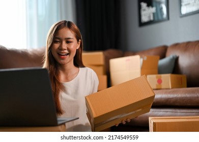 Charming Millennial Asian SME Business Owner Updating Her Stock And Shipping Status On Laptop Computer While Preparing A Shipping Boxes In The Living Room.