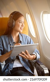Charming millennial asian woman passenger at her window seat with her wireless headphones using portable tablet and looking out the plane's window.